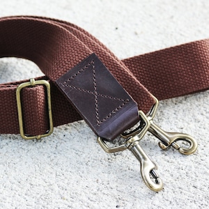 May include: A brown canvas camera strap with a leather patch and brass hardware. The strap has two swivel hooks.