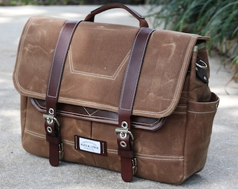 May include: A brown waxed canvas messenger bag with leather accents. The bag features a flap closure, a top handle, and adjustable leather straps with silver buckles. The bag has a small rectangular tag that reads "ALEX M. LYNCH".