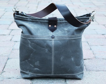 May include: A gray canvas tote bag with a brown leather strap and accents. The bag has a front pocket and a leather closure with copper rivets. The bag is sitting on a brick surface.
