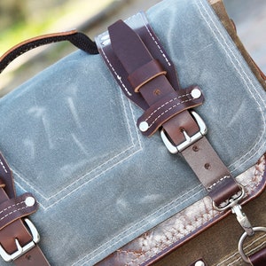 May include: A gray canvas backpack with brown leather straps and buckles. The backpack has a top handle and a front pocket.
