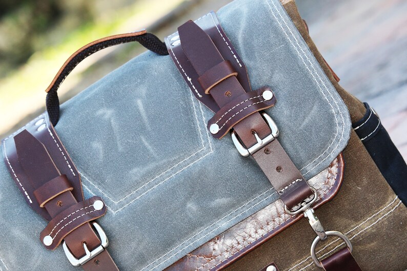 May include: A gray canvas backpack with brown leather straps and buckles. The backpack has a top handle and a front pocket.