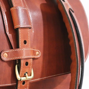 May include: Close-up of a brown leather satchel bag with a brass buckle and white stitching. The bag has a black leather strap.