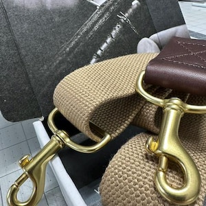 Puede incluir: Una correa de lona beige con un acento de cuero marrón y cierres de metal dorado. La correa es para un bolso o una cartera. La correa está hecha a mano por Alex M Lynch.