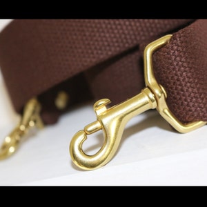 May include: A dark brown woven fabric strap featuring two polished gold-tone metal swivel lobster claw clasps attached to the ends. The strap has a textured, durable appearance with a thick weave, and the metal hardware has a smooth, reflective finish.