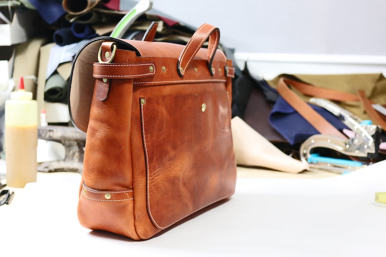 May include: A brown leather satchel bag with a flap closure and a brass buckle. The bag has a small pocket on the front and a strap for carrying.