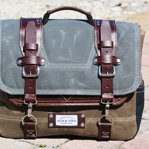 May include: A brown leather and canvas messenger bag with two large buckles and a smaller buckle on the front. The bag has a brown leather handle and a label that reads "HANDCRAFTED GOODS ALEX M. LYNCH MADE IN USA".