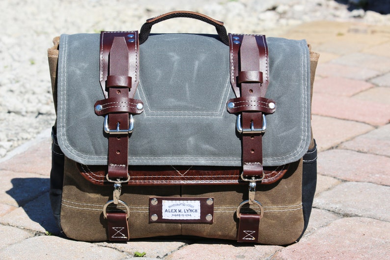 May include: A brown leather and canvas messenger bag with two large buckles and a smaller buckle on the front. The bag has a brown leather handle and a label that reads "HANDCRAFTED GOODS ALEX M. LYNCH MADE IN USA".