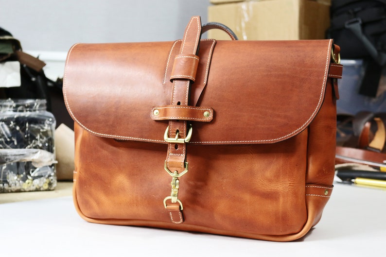 May include: A brown leather messenger bag with a brass buckle and clasp. The bag has a flap closure and a shoulder strap.