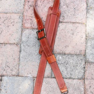 May include: A brown leather camera strap with a metal buckle and two metal clips. The strap is crossed and laid flat on a brick surface.