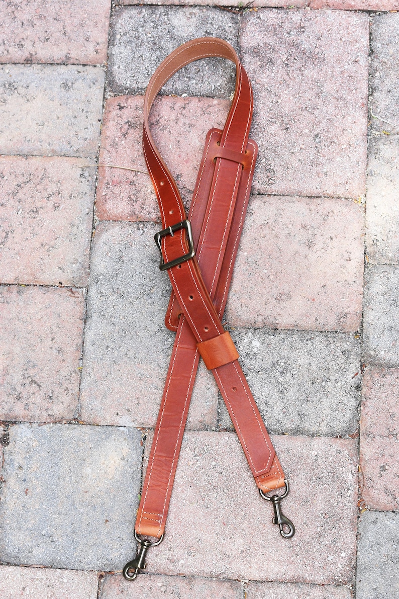 May include: A brown leather camera strap with a metal buckle and two metal clips. The strap is crossed and laid flat on a brick surface.