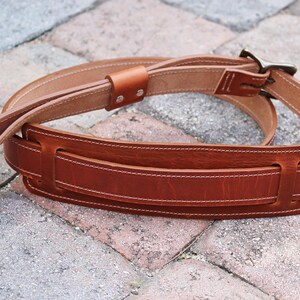 May include: A brown leather guitar strap with a stitched design and a metal buckle. The strap is folded in half and lying on a brick surface.