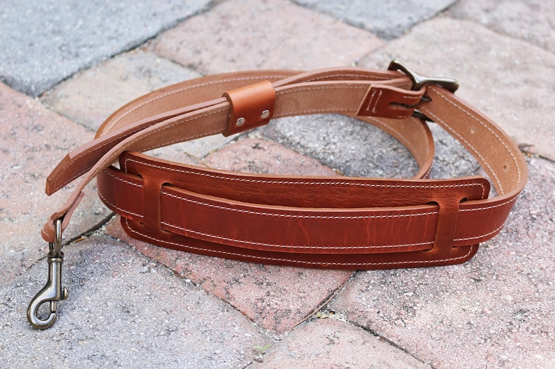 May include: A brown leather guitar strap with a stitched design and a metal buckle. The strap is folded in half and lying on a brick surface.