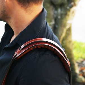 May include: A brown leather camera strap with a padded shoulder rest. The strap is worn over a person's shoulder.