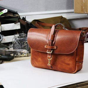 May include: A brown leather messenger bag with a brass buckle and a strap. The bag is sitting on a white surface.