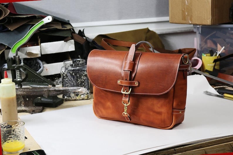 May include: A brown leather messenger bag with a brass buckle and a strap. The bag is sitting on a white surface.