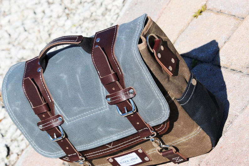 May include: A brown and gray canvas messenger bag with leather straps and buckles. The bag has a flap closure and a small pocket on the front. The bag is made by "Alex & Sons".