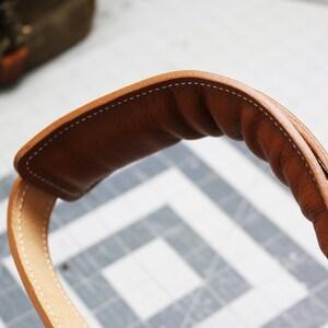 May include: Close-up of a brown leather strap with white stitching. The strap is folded over and has a rounded shape.