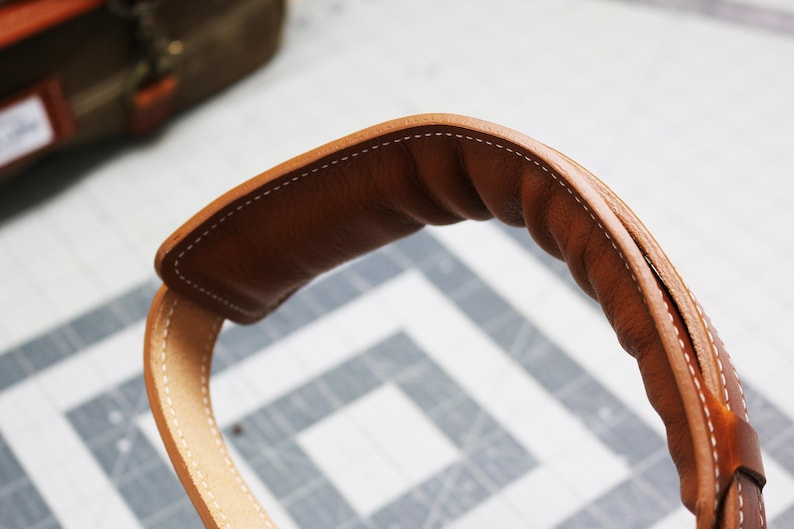 May include: Close-up of a brown leather strap with white stitching. The strap is folded over and has a rounded shape.