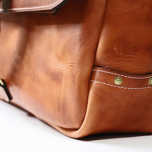 May include: A brown leather satchel bag with a brass buckle and a flap closure. The bag has a white stitched detail and brass rivets on the bottom.