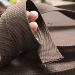 May include: Close-up of a roll of brown canvas webbing. The webbing is a solid, dark brown color with a textured weave. A hand holds a section of the webbing, showing its flexibility and detail. The webbing is likely used for straps or belts.