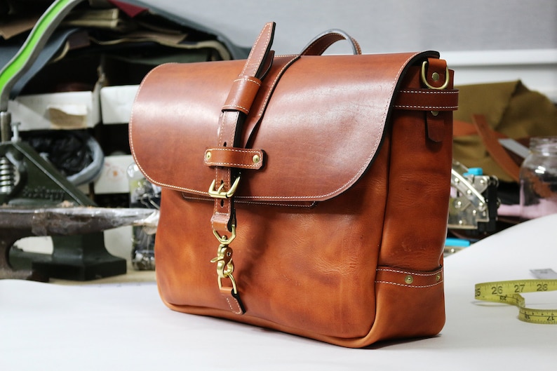May include: A brown leather messenger bag with a brass buckle and strap. The bag has a flap closure and a shoulder strap.