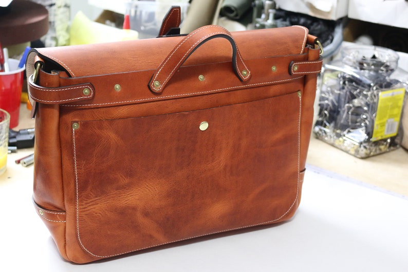 May include: A brown leather satchel bag with a flap closure and a brass buckle. The bag has a front pocket and a white stitching detail.