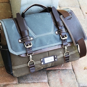 May include: A brown leather messenger bag with a blue canvas flap and a brown leather strap. The bag has a metal buckle and a metal ring. The bag has a label that says "ALOUD SUPPLY CO."