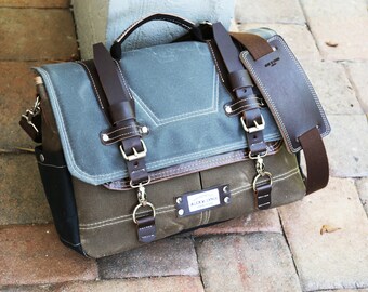 May include: A brown leather messenger bag with a blue canvas flap and a brown leather strap. The bag has a metal buckle and a metal ring. The bag has a label that says "ALOUD SUPPLY CO."