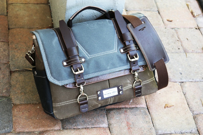 May include: A brown leather messenger bag with a blue canvas flap and a brown leather strap. The bag has a metal buckle and a metal ring. The bag has a label that says "ALOUD SUPPLY CO."