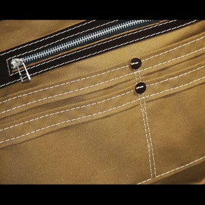 May include: Close-up of a brown canvas bag with a zippered pocket and two brown buttons. The bag has white stitching around the edges.