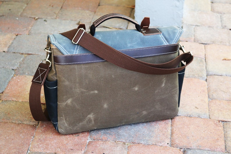 May include: A brown canvas messenger bag with a brown leather handle and strap. The bag has a blue canvas flap with brown leather trim.