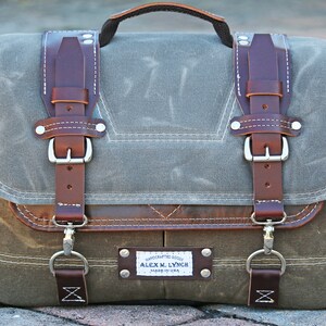May include: A brown leather and canvas messenger bag with a metal buckle closure and a leather strap. The bag has a brown leather patch with the text "HANDCRAFTED GOODS ALEX M. LYNCH MADE IN USA".