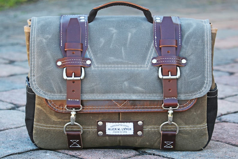 May include: A brown leather and canvas messenger bag with a metal buckle closure and a leather strap. The bag has a brown leather patch with the text "HANDCRAFTED GOODS ALEX M. LYNCH MADE IN USA".