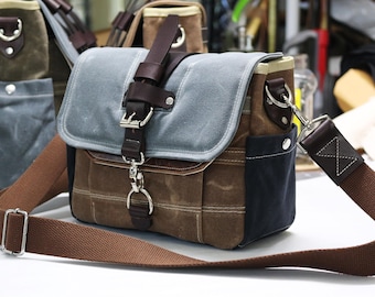 Puede incluir: Bolso mensajero de lona con solapa gris y detalles en cuero marrón. El bolso tiene una correa de hombro marrón y herrajes plateados. Cuenta con un bolsillo frontal y cierre de hebilla. Se ven otros bolsos similares en el fondo.