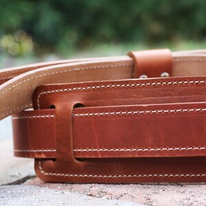 May include: A brown leather belt with white stitching. The belt is made of thick, durable leather and has a simple, classic design.