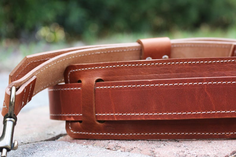 May include: A brown leather belt with white stitching. The belt is made of thick, durable leather and has a simple, classic design.