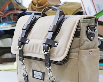 Puede incluir: Un bolso de mensajero de lona beige con correas y hebillas de cuero marrón. El bolso tiene un cierre de solapa y un bolsillo frontal. El bolso tiene una hebilla de metal y una etiqueta de metal con el texto "Alec & Lynch" en él.