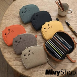 Puede incluir: Una colección de siete monederos y tarjeteros pequeños con forma de gato en colores naranja, gris, beige, amarillo, negro y azul. Cada uno tiene una superficie texturizada, dos ojos redondos y bigotes. Un monedero amarillo está abierto, mostrando varias ranuras para tarjetas.