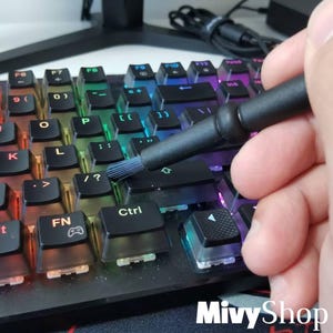 Puede incluir: Un cepillo de limpieza negro con cerdas suaves que se utiliza para limpiar un teclado mecánico con teclas negras y retroiluminación RGB multicolor. El teclado incluye teclas de función, una tecla Ctrl y un icono de juego en la tecla FN.