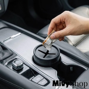 Puede incluir: Una papelera cilíndrica negra para coche con una tapa de silicona flexible con ranura en cruz, diseñada para encajar en el portavasos de un vehículo. El recipiente es de material mate y se muestra siendo usado para desechar un papel arrugado.
