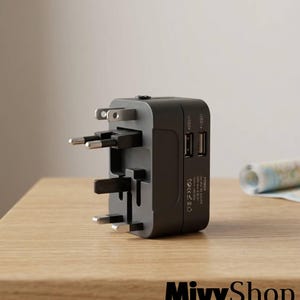 Pode incluir: Um adaptador de viagem universal cinza-escuro com vários pinos retráteis de um lado e duas portas USB no painel lateral. O dispositivo inclui especificações técnicas impressas e símbolos regulatórios perto das portas USB.