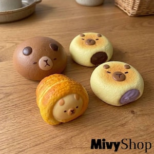 May include: Four small, round, bear-shaped food items. One is a smooth brown bun, one is a light yellow bun with a dark brown filling, one is a light yellow bun with a purple filling, and one is a golden-brown, textured, rolled pastry shape.