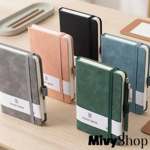 Puede incluir: Una colección de cinco libretas de bolsillo en negro, gris, melocotón, verde y azul. Cada libreta presenta una cubierta texturizada, una banda de papel blanca con el texto POCKET BOOK, una cinta elástica vertical y un soporte para bolígrafo.
