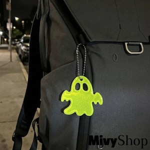 Puede incluir: Un llavero reflectante de color amarillo neón con forma de fantasma, sujeto a una mochila negra mediante una cadena de bolas de metal plateado. La figura del fantasma presenta dos aberturas ovaladas para los ojos y un borde inferior ondulado.