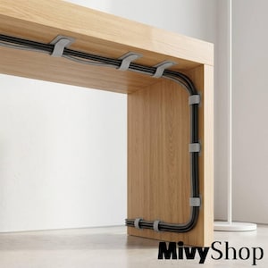 Puede incluir: Un escritorio de madera de roble claro con una serie de clips de plástico gris para cables fijados en la parte inferior y el panel lateral. Los clips aseguran un cable de alimentación trenzado negro a lo largo del marco de madera.
