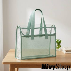 May include: A rectangular tote bag featuring a clear plastic upper section and a sage green mesh lower section. The bag includes two matching sage green fabric handles and a top flap secured with two small white snap buttons for closure.
