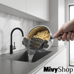 Puede incluir: Un colador de silicona gris oscuro con clip, sujeto al borde de una cacerola de acero inoxidable llena de pasta rotini cocida. El colador presenta un diseño curvo y perforado con clips laterales para drenar el agua en el fregadero.