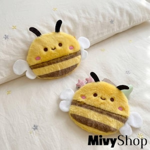 May include: Two plush bee-shaped pouches with soft yellow and brown striped fabric, white felt wings, and embroidered faces featuring small dark eyes and pink cheeks. The items have black antennae and are positioned on a light fabric surface.