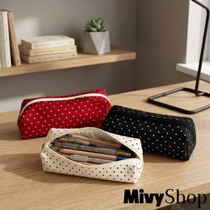Op de afbeelding: Drie rechthoekige etuis met een wit stippenpatroon op rode, zwarte en crèmekleurige stof. Elk etui heeft een ritssluiting. Het crèmekleurige etui is geopend en toont verschillende houten potloden en metalen pennen aan de binnenkant.