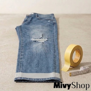 May include: Blue denim jeans featuring a distressed knee area and a light gray ribbon trim applied to the bottom hem. A roll of yellow masking tape and a white measuring tape showing measurements in inches are positioned next to the jeans.